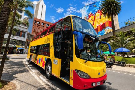Bus panorámico de dos pisos en el centro de Medellín, con palmas y un mural de colores al fondo